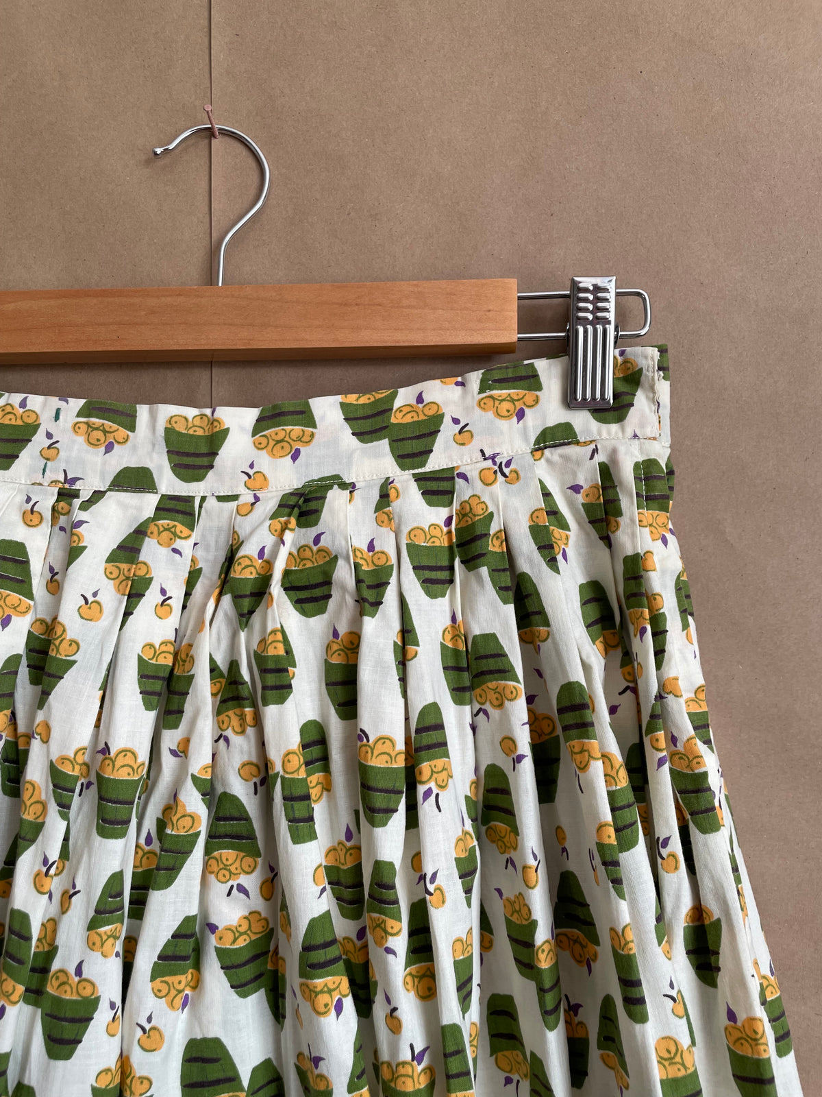 A vintage handmade pleated midi skirt with an apple basket novelty print in olive, mustard, and cream, shown against a kraft background