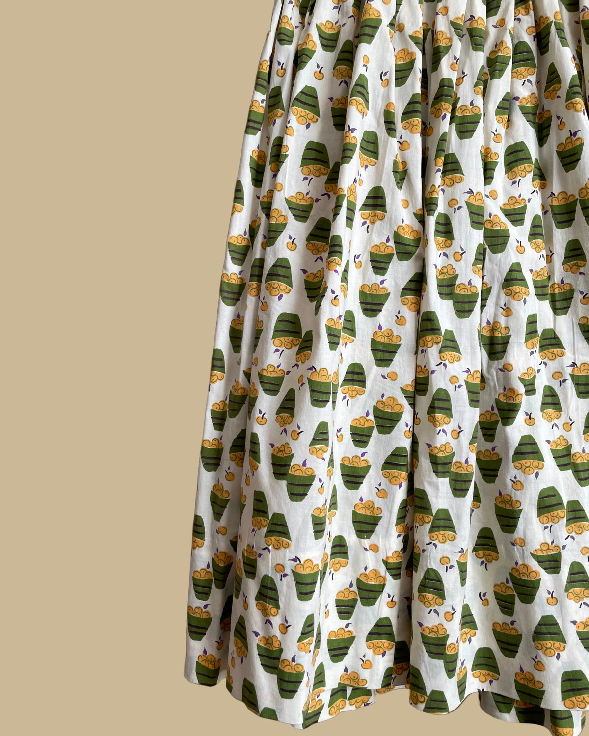 A vintage handmade pleated midi skirt with an apple basket novelty print in olive, mustard, and cream, shown against a kraft background