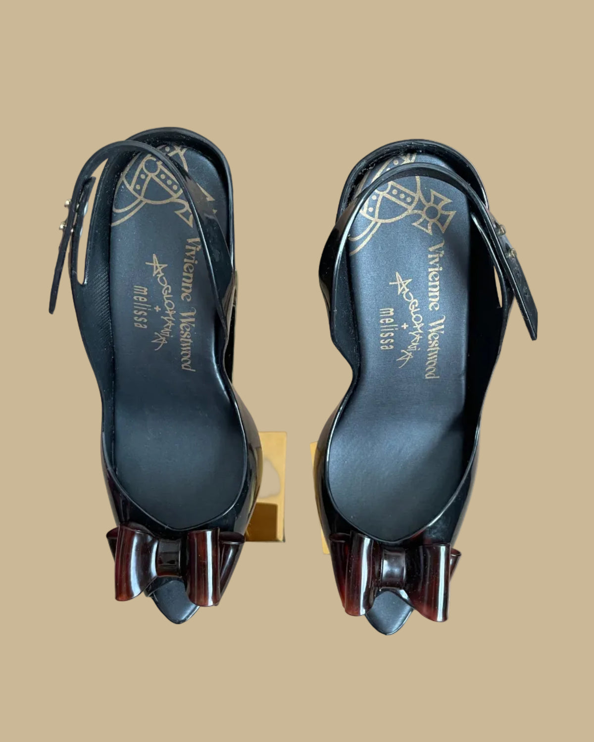 A pair of black jelly slingback heels from the Vivienne Westwood Anglomania x Melissa collaboration with a tortoiseshell bow, displayed on gold stands against a kraft background