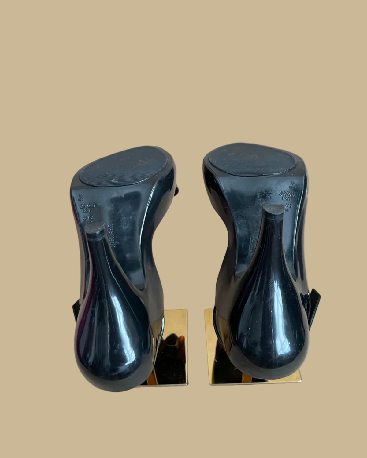 A pair of black jelly slingback heels from the Vivienne Westwood Anglomania x Melissa collaboration with a tortoiseshell bow, displayed on gold stands against a kraft background
