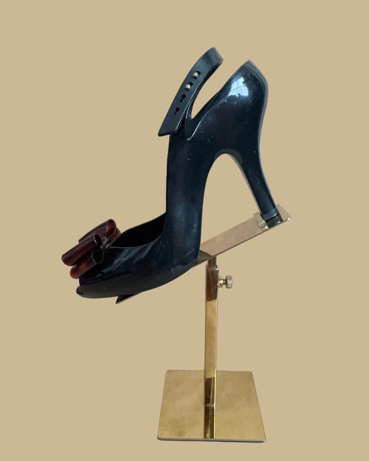 A pair of black jelly slingback heels from the Vivienne Westwood Anglomania x Melissa collaboration with a tortoiseshell bow, displayed on gold stands against a kraft background