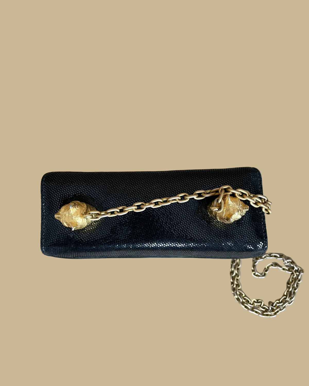A small black textured handbag with a gold-tone chain strap and decorative gold-tone hardware, photographed against a neutral beige backdrop.