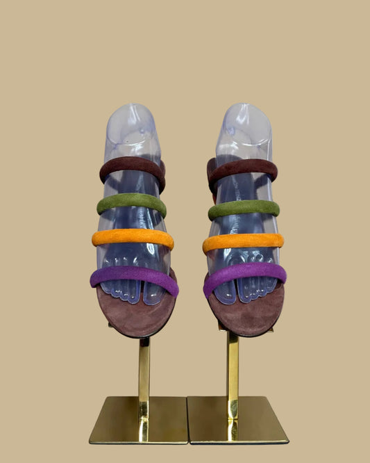 A pair of multi-strap suede wedge sandals in brown, olive, mustard, and purple, shown on clear display forms against a neutral beige background
