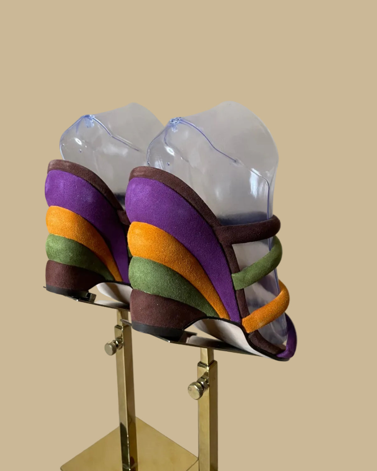 A pair of multi-strap suede wedge sandals in brown, olive, mustard, and purple, shown on clear display forms against a neutral beige background