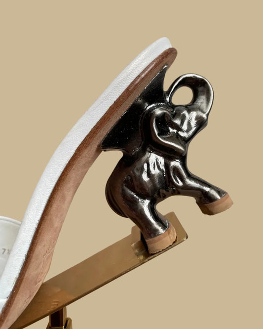 A pair of white leather Ramón Tenza slide sandals with a sculptural gunmetal elephant-shaped heel, displayed on gold stands against a kraft background