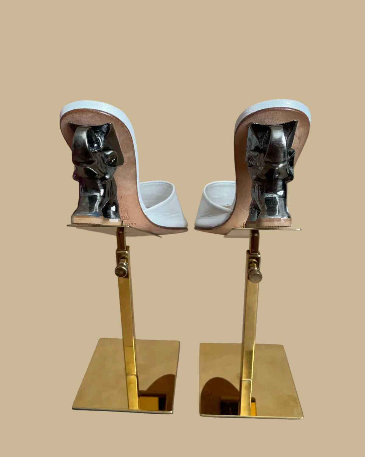 A pair of white leather Ramón Tenza slide sandals with a sculptural gunmetal elephant-shaped heel, displayed on gold stands against a kraft background