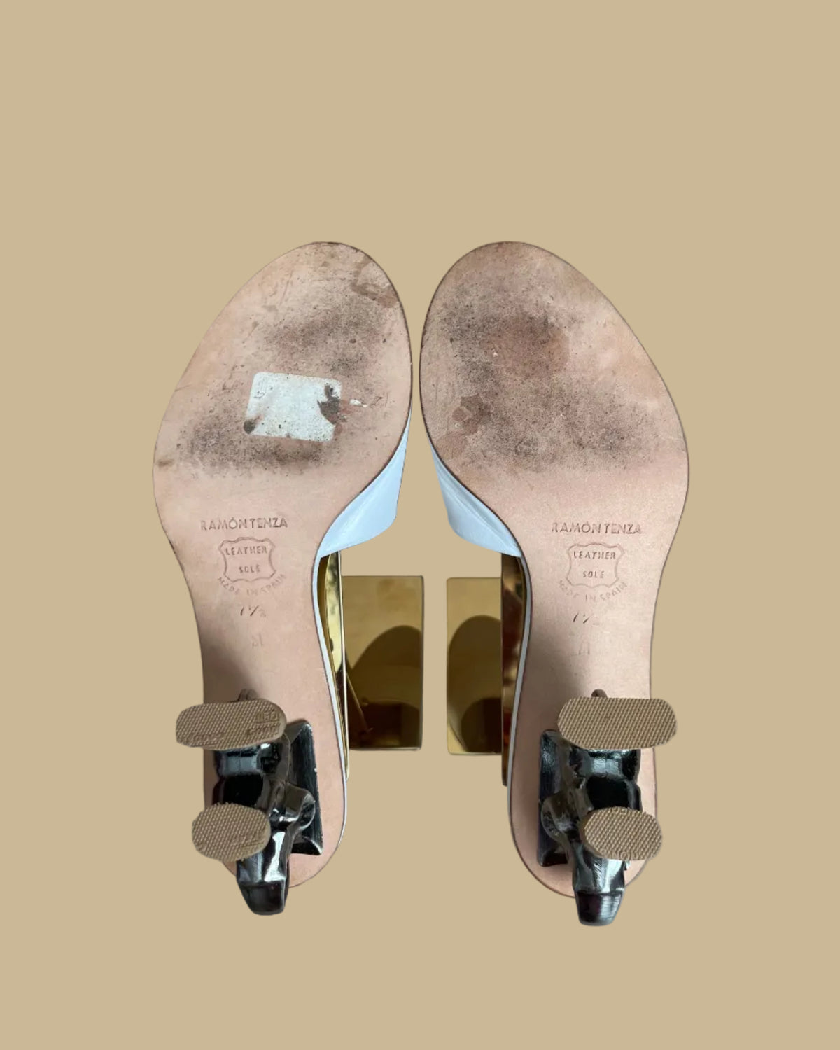A pair of white leather Ramón Tenza slide sandals with a sculptural gunmetal elephant-shaped heel, displayed on gold stands against a kraft background