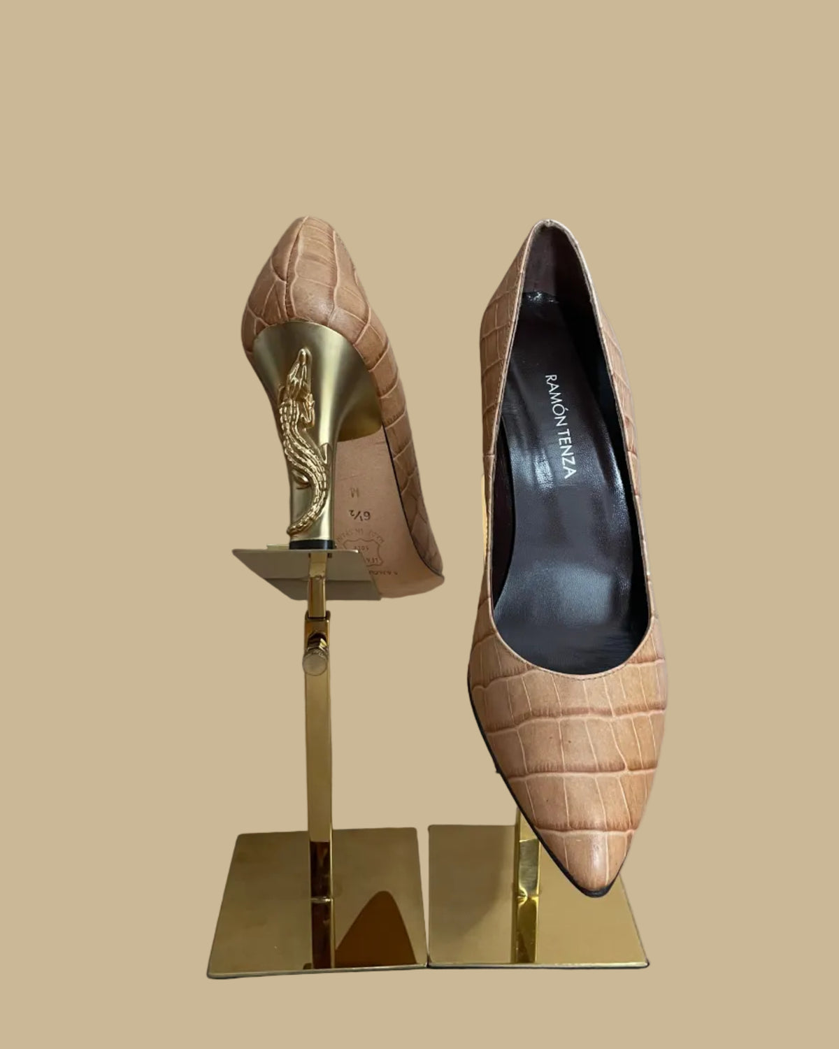 A pair of pointed-toe high heels in a tan crocodile-embossed leather, displayed on a gold-colored stand against a neutral beige background. The brand name Ramon Tenza is visible on the black insoles.