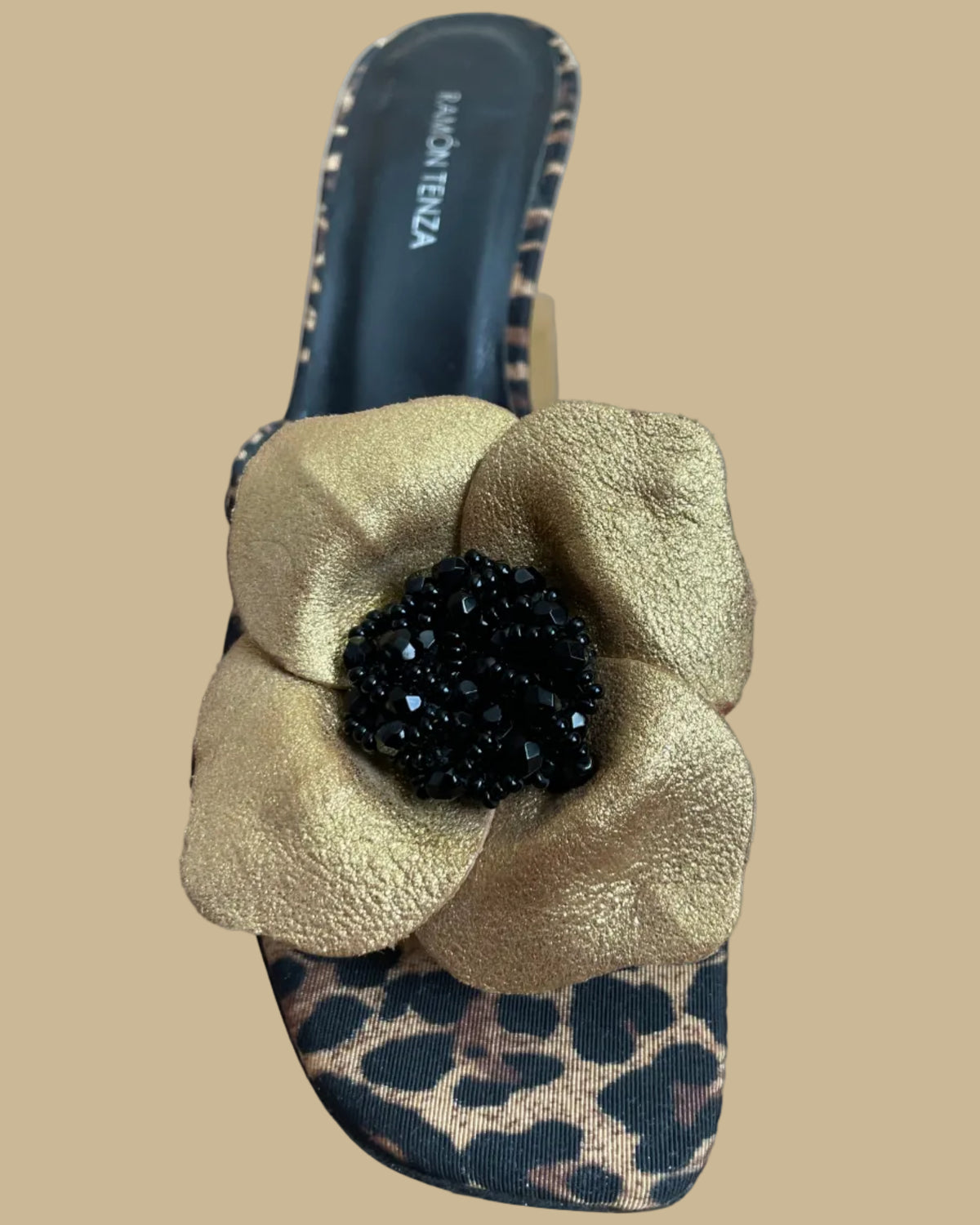 A pair of leopard print Ramón Tenza slingback sandals with a gold leather flower and black beaded center, displayed on gold stands against a kraft background