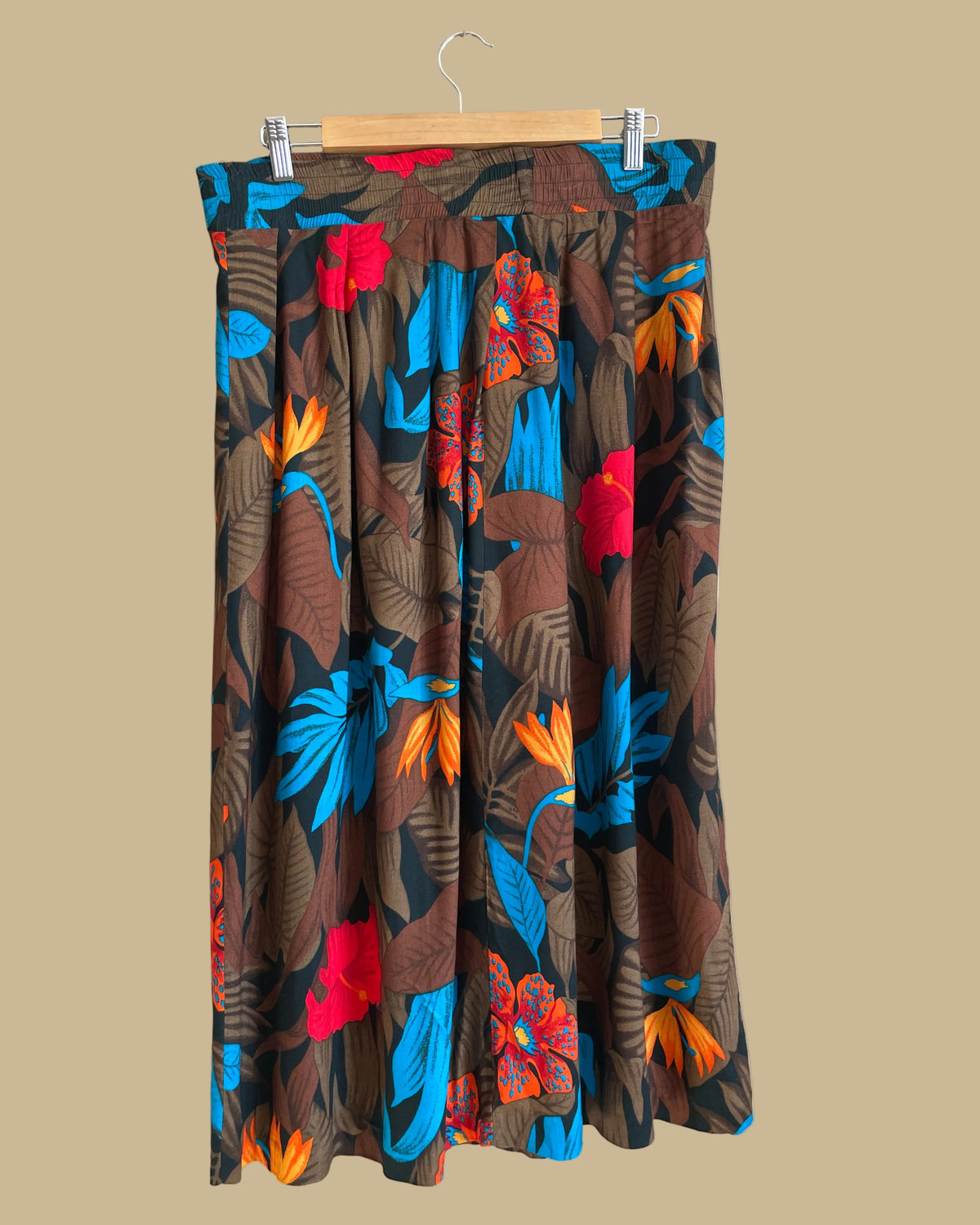 A vintage tropical bird-of-paradise print pleated skirt in brown, black, teal, and orange tones, shown against a kraft background