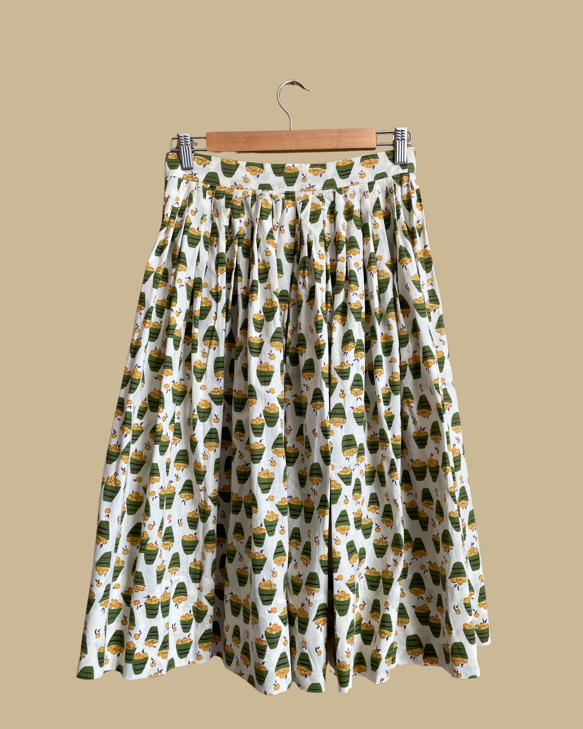 A vintage handmade pleated midi skirt with an apple basket novelty print in olive, mustard, and cream, shown against a kraft background