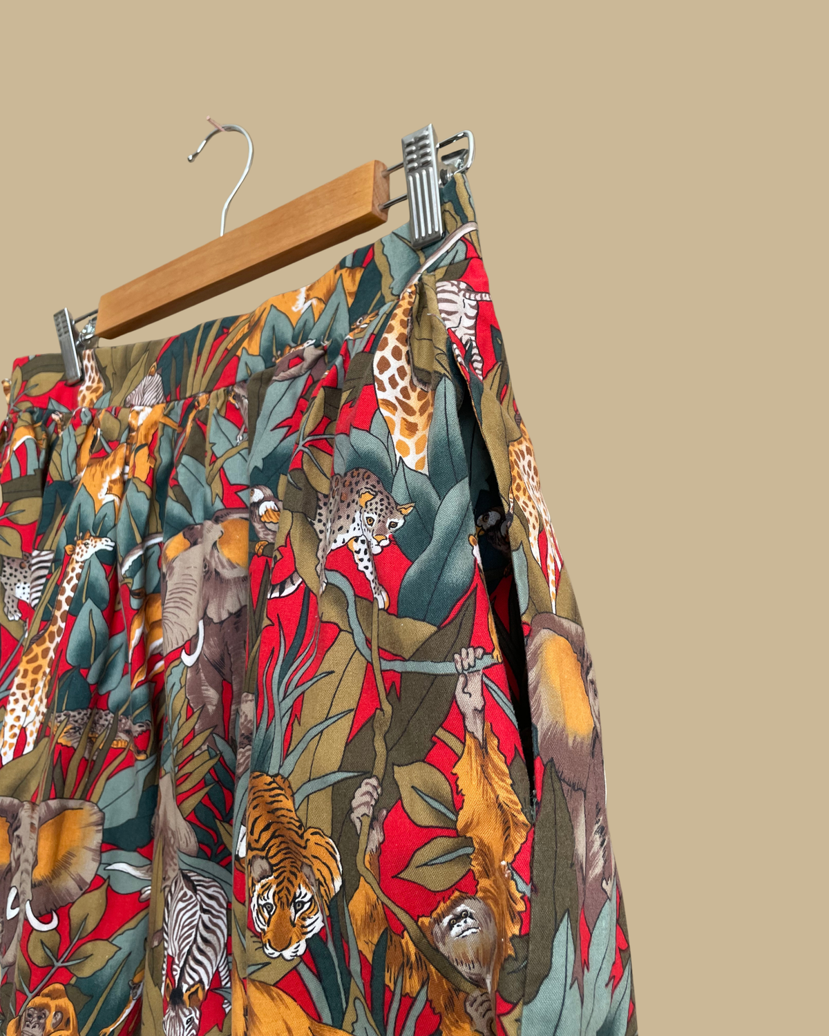A vintage handmade pleated midi skirt with a jungle safari animal print featuring elephants, giraffes, zebras, and tigers in red, olive, and teal, shown against a kraft background