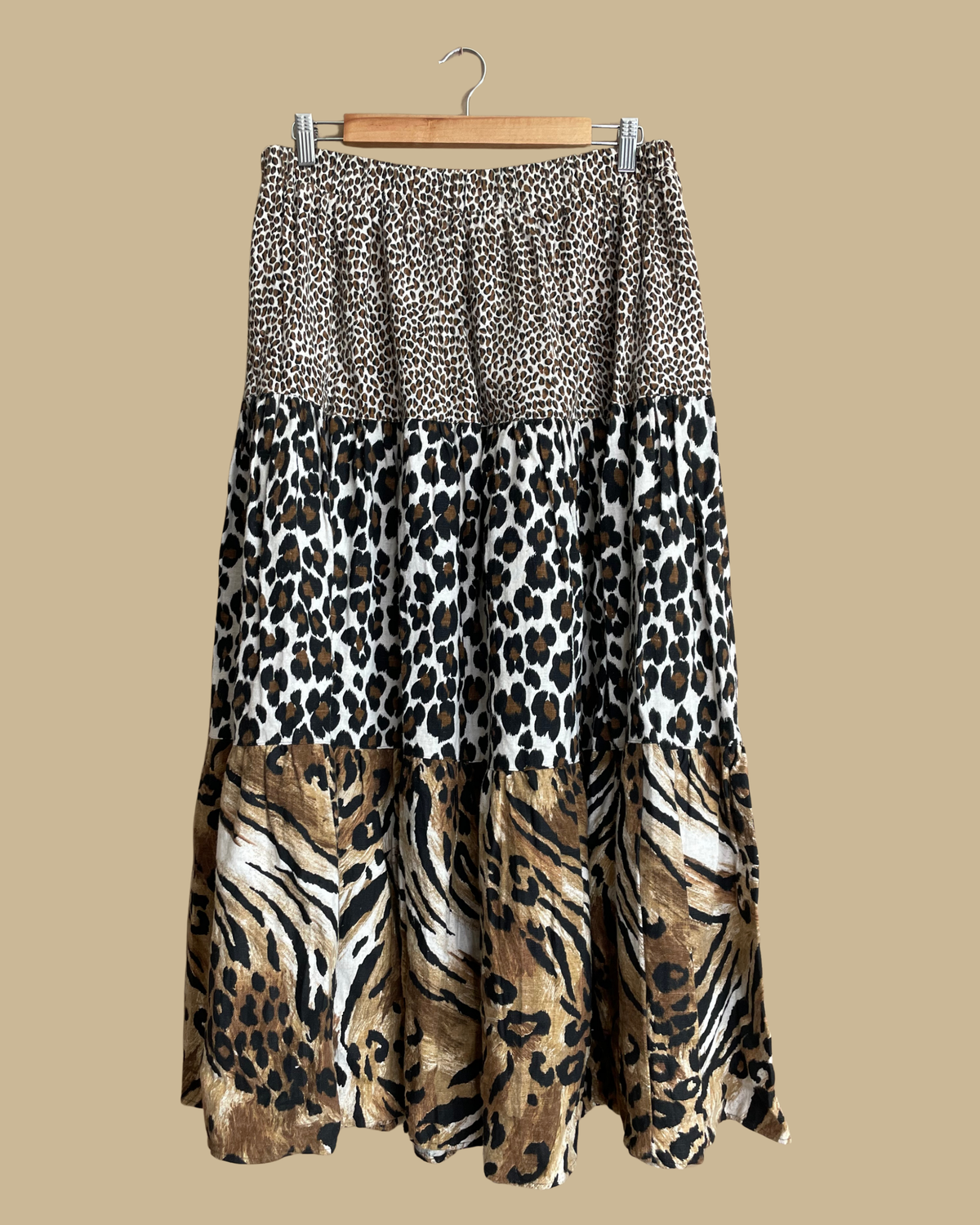 A tiered maxi skirt with mixed animal prints, leopard spots at the waist transitioning to a tiger and zebra-inspired border at the hem, on a hanger against a neutral beige background