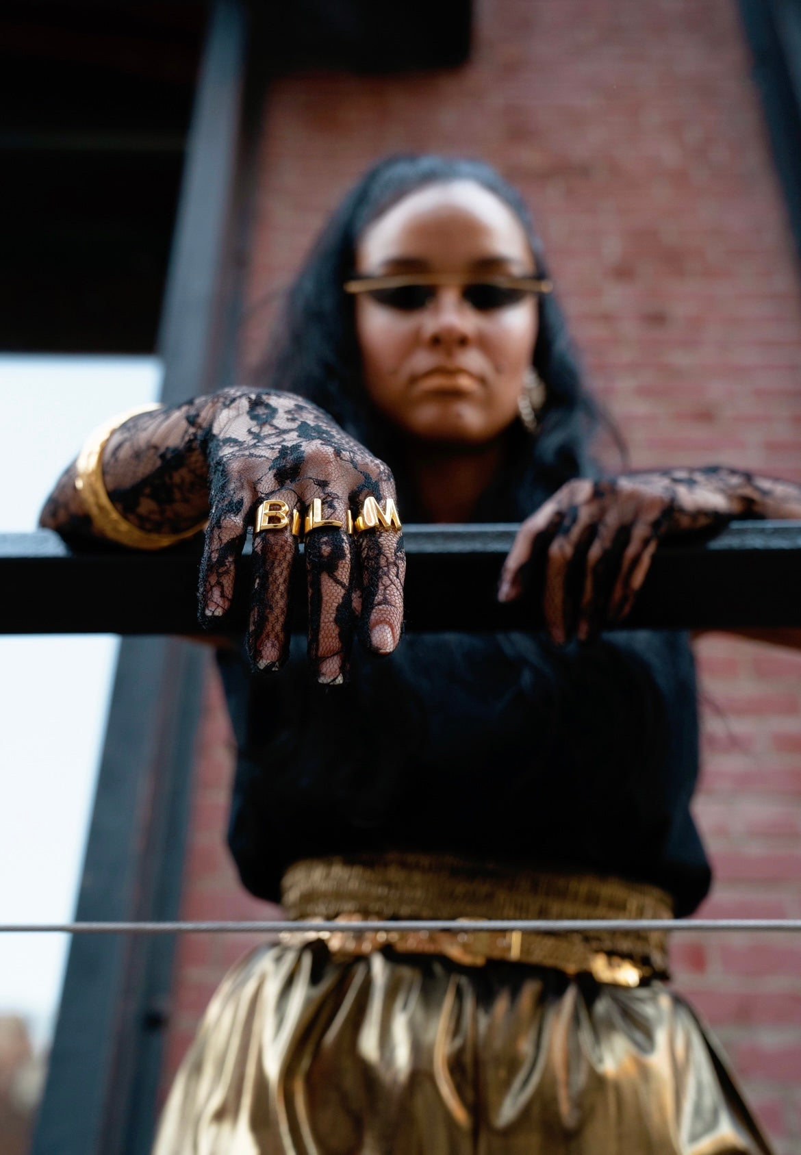 A person wearing three golden-colored rings with the letters 'B', 'L', and 'M' on their fingers, standing by a railing with their hands and feet adorned with black and gold decorative patterns.