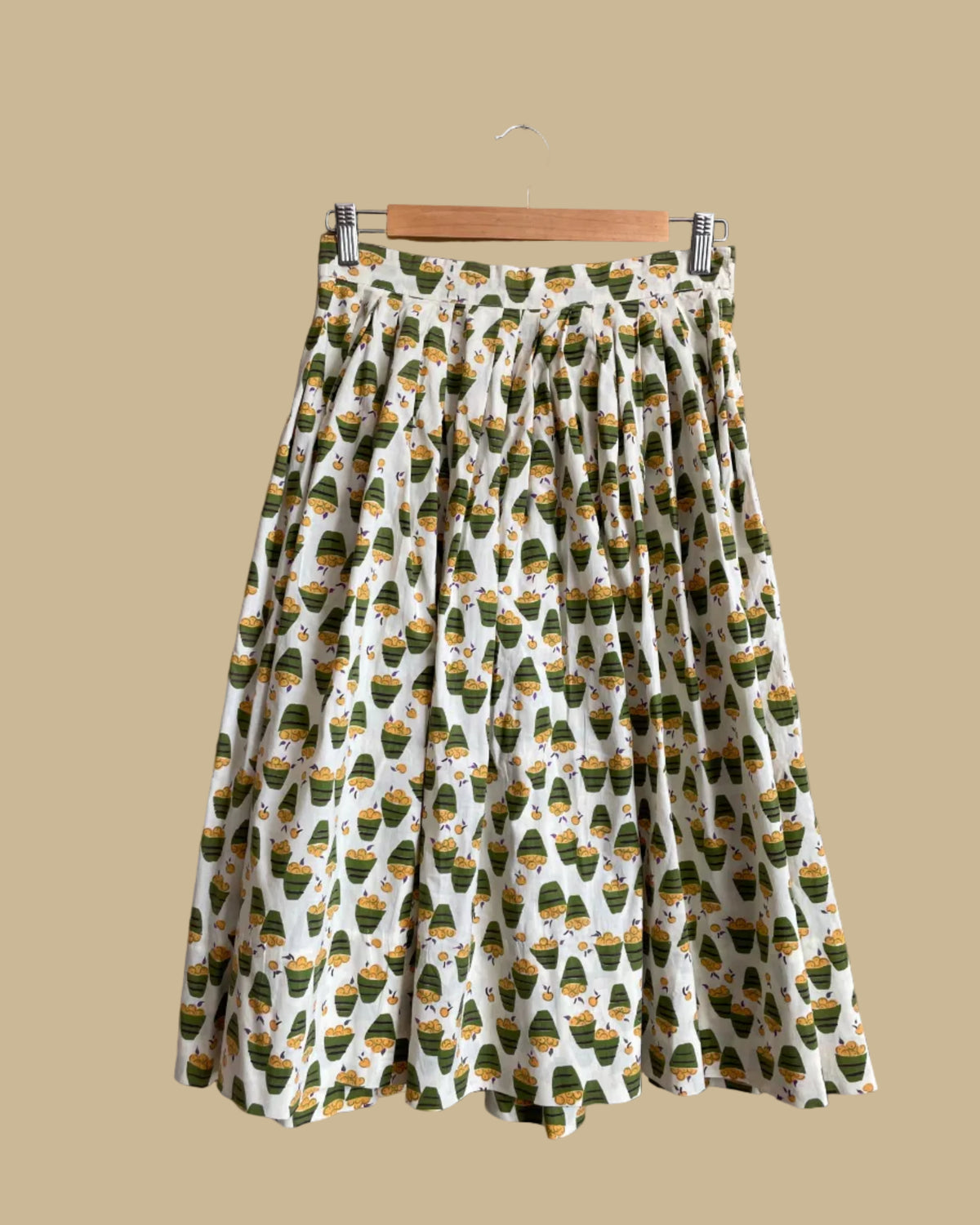 A vintage handmade pleated midi skirt with an apple basket novelty print in olive, mustard, and cream, shown against a kraft background