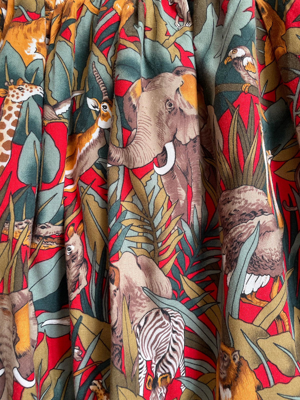 A vintage handmade pleated midi skirt with a jungle safari animal print featuring elephants, giraffes, zebras, and tigers in red, olive, and teal, shown against a kraft background