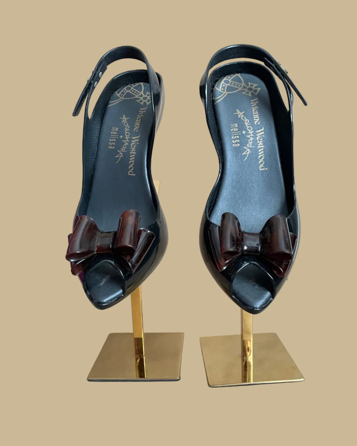 A pair of black jelly slingback heels from the Vivienne Westwood Anglomania x Melissa collaboration with a tortoiseshell bow, displayed on gold stands against a kraft background