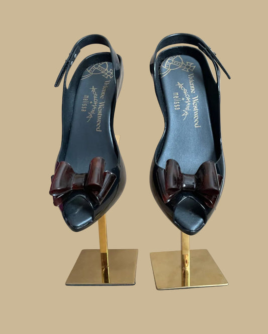 A pair of black jelly slingback heels from the Vivienne Westwood Anglomania x Melissa collaboration with a tortoiseshell bow, displayed on gold stands against a kraft background