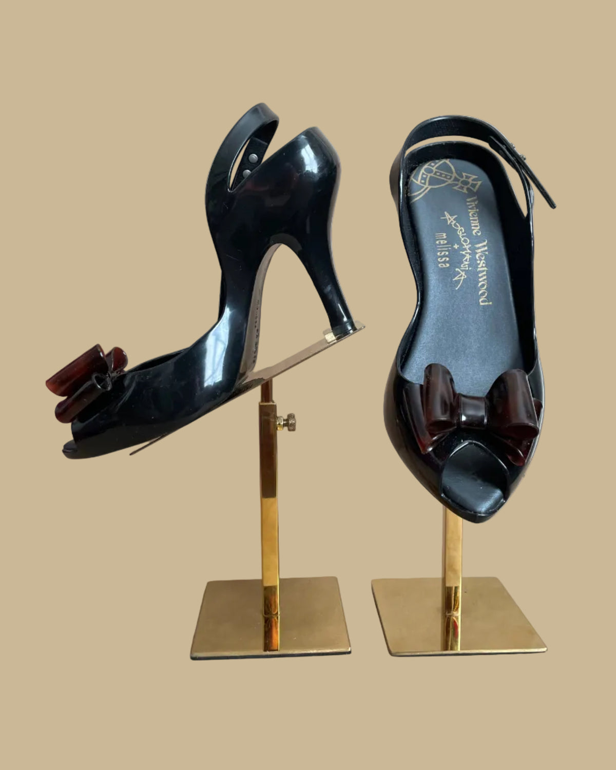 A pair of black jelly slingback heels from the Vivienne Westwood Anglomania x Melissa collaboration with a tortoiseshell bow, displayed on gold stands against a kraft background