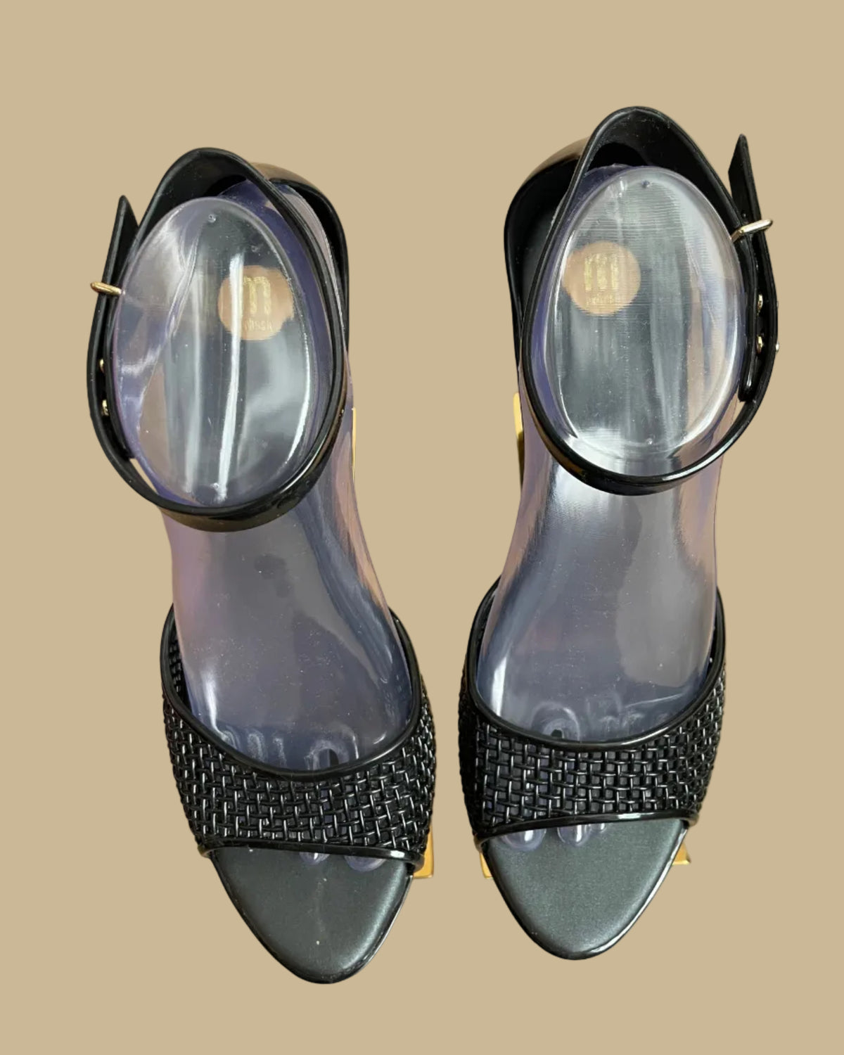 A pair of black Melissa jelly wedge sandals with a woven-texture toe strap and ankle buckle, displayed on gold stands against a kraft background