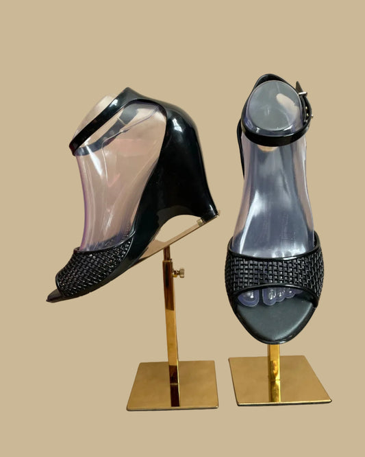 A pair of black Melissa jelly wedge sandals with a woven-texture toe strap and ankle buckle, displayed on gold stands against a kraft background
