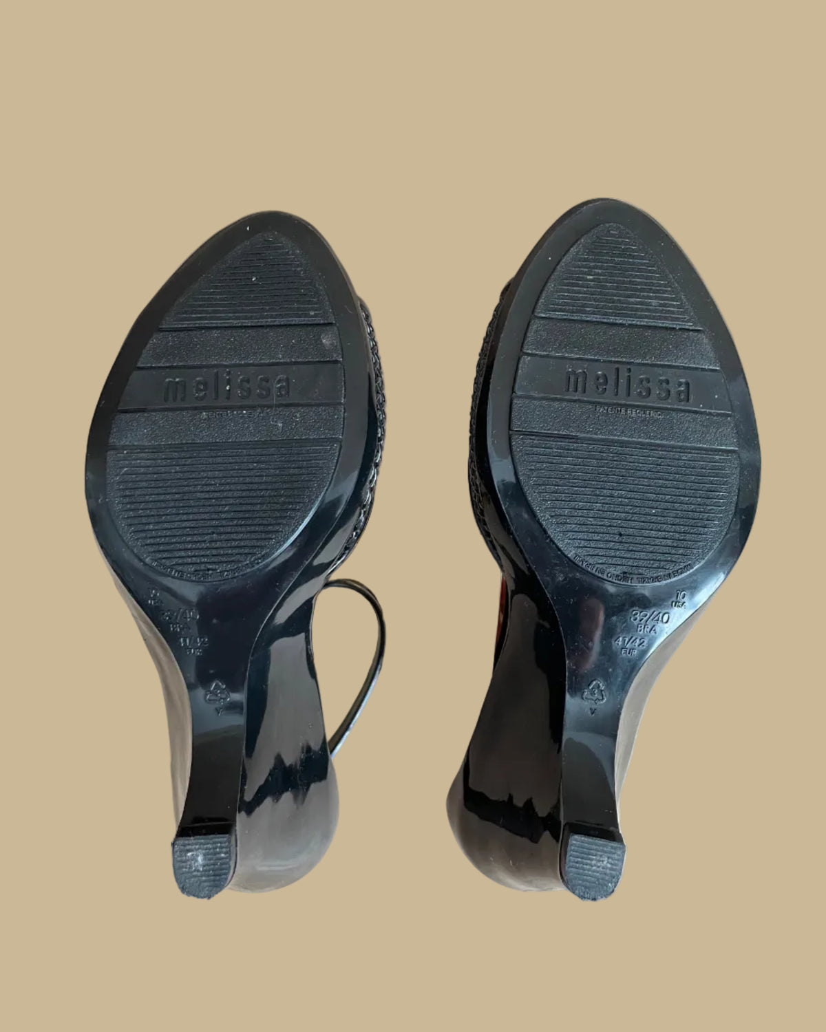 A pair of black Melissa jelly wedge sandals with a woven-texture toe strap and ankle buckle, displayed on gold stands against a kraft background