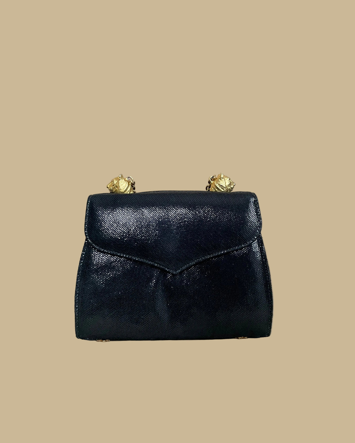 A small black textured handbag with a gold-tone chain strap and decorative gold-tone hardware, photographed against a neutral beige backdrop.