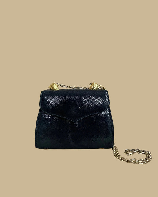A small black textured handbag with a gold-tone chain strap and decorative gold-tone hardware, photographed against a neutral beige backdrop.