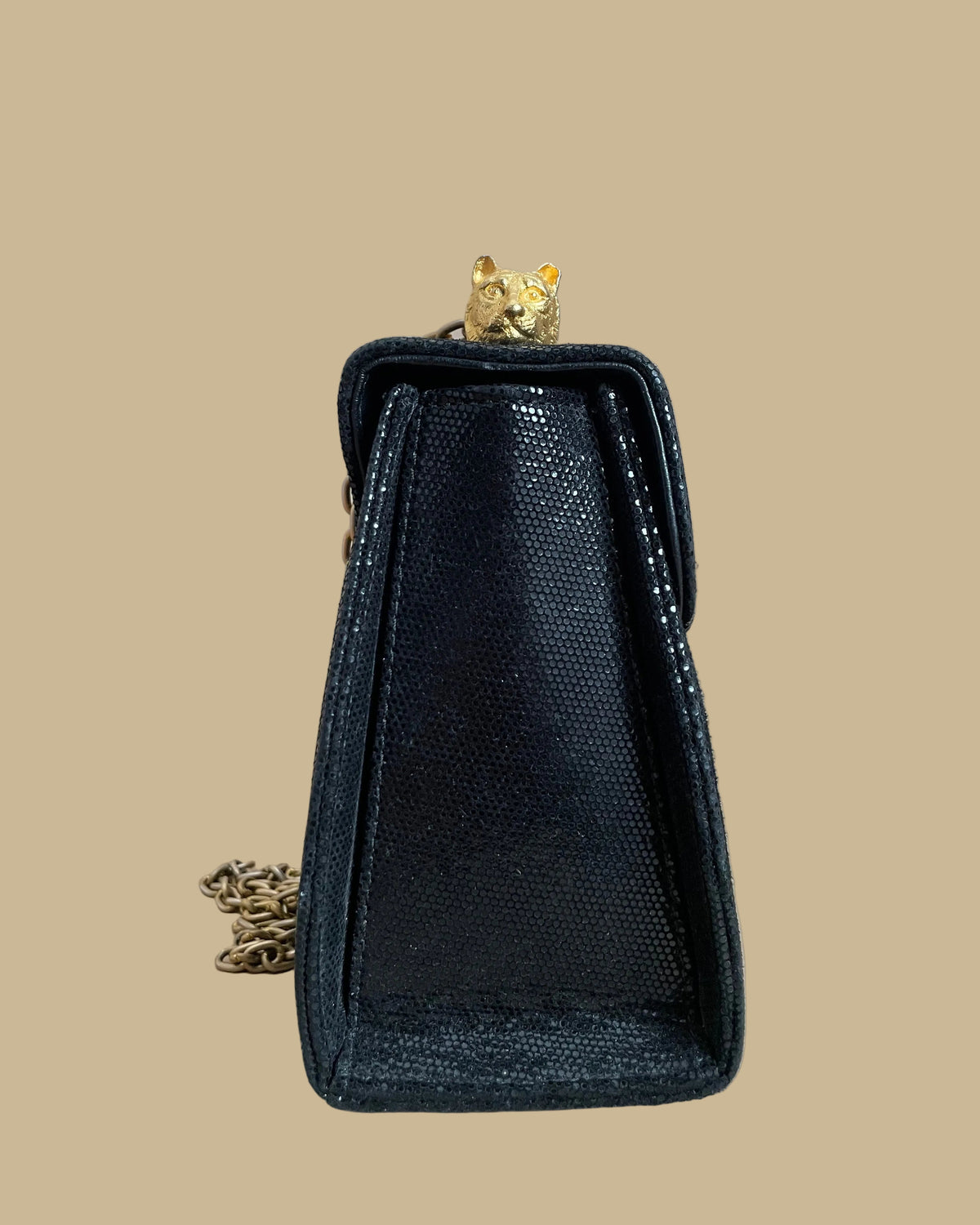 A small black textured handbag with a gold-tone chain strap and decorative gold-tone hardware, photographed against a neutral beige backdrop.