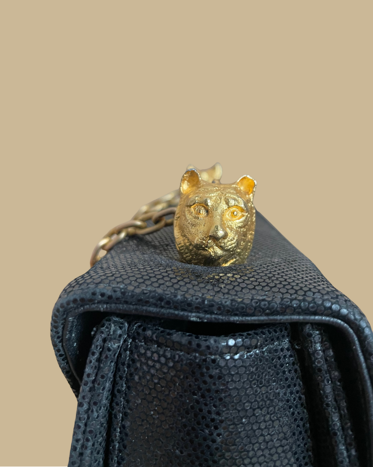 A close-up of a textured black handbag featuring a gold tiger head charm resting on top, with a gold chain strap visible against a solid beige background.