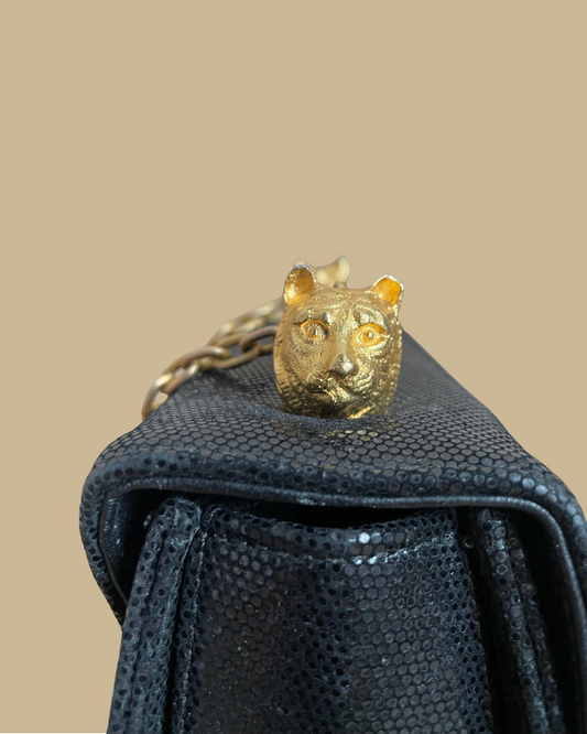 A close-up of a textured black handbag featuring a gold tiger head charm resting on top, with a gold chain strap visible against a solid beige background.