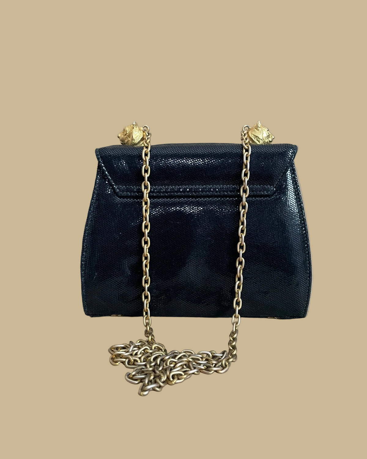 A small black textured handbag with a gold-tone chain strap and decorative gold-tone hardware, photographed against a neutral beige backdrop.