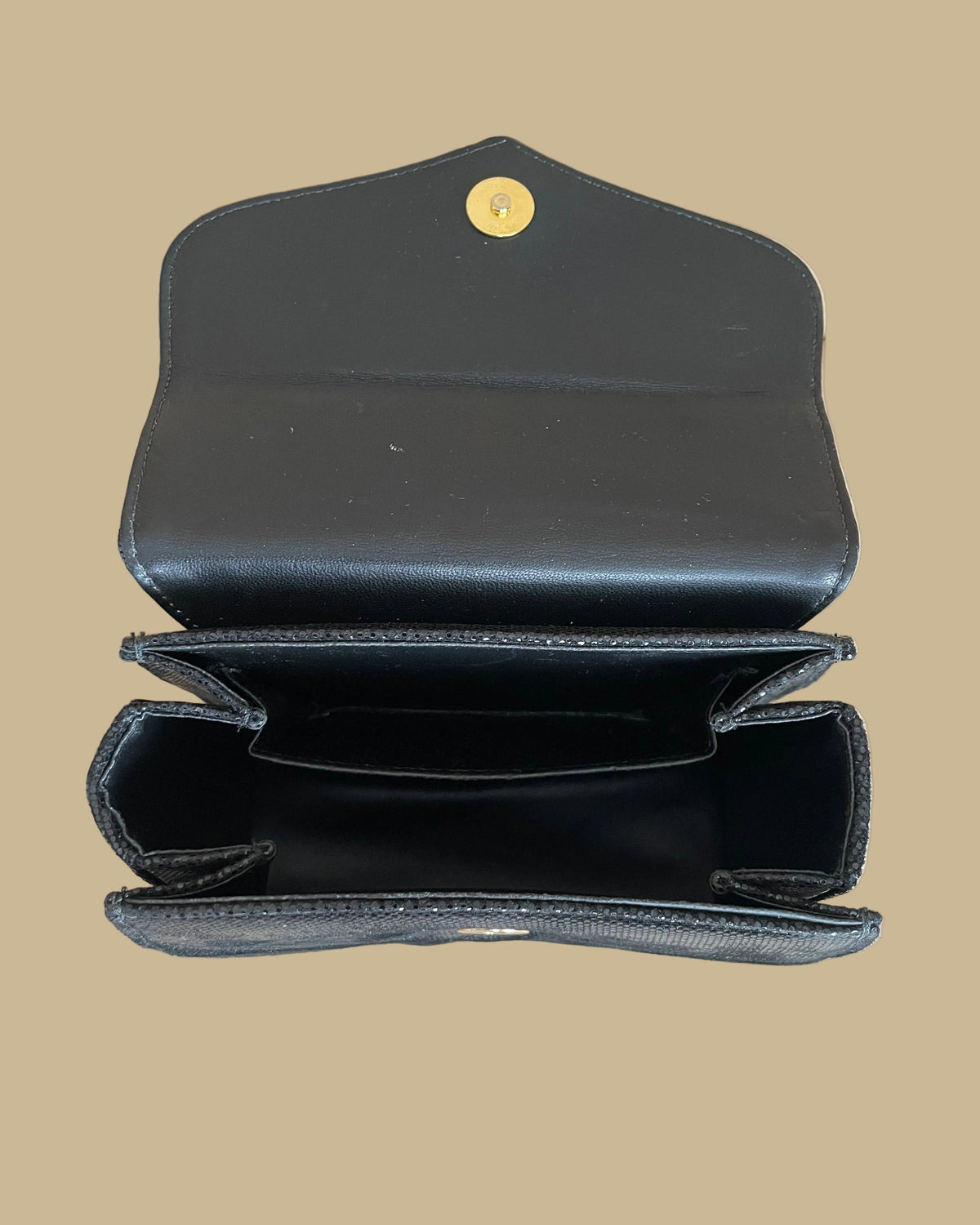 A small black textured handbag with a gold-tone chain strap and decorative gold-tone hardware, photographed against a neutral beige backdrop.