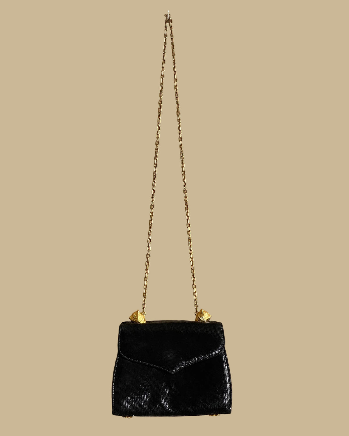 A small black textured handbag with a gold-tone chain strap and decorative gold-tone hardware, photographed against a neutral beige backdrop.