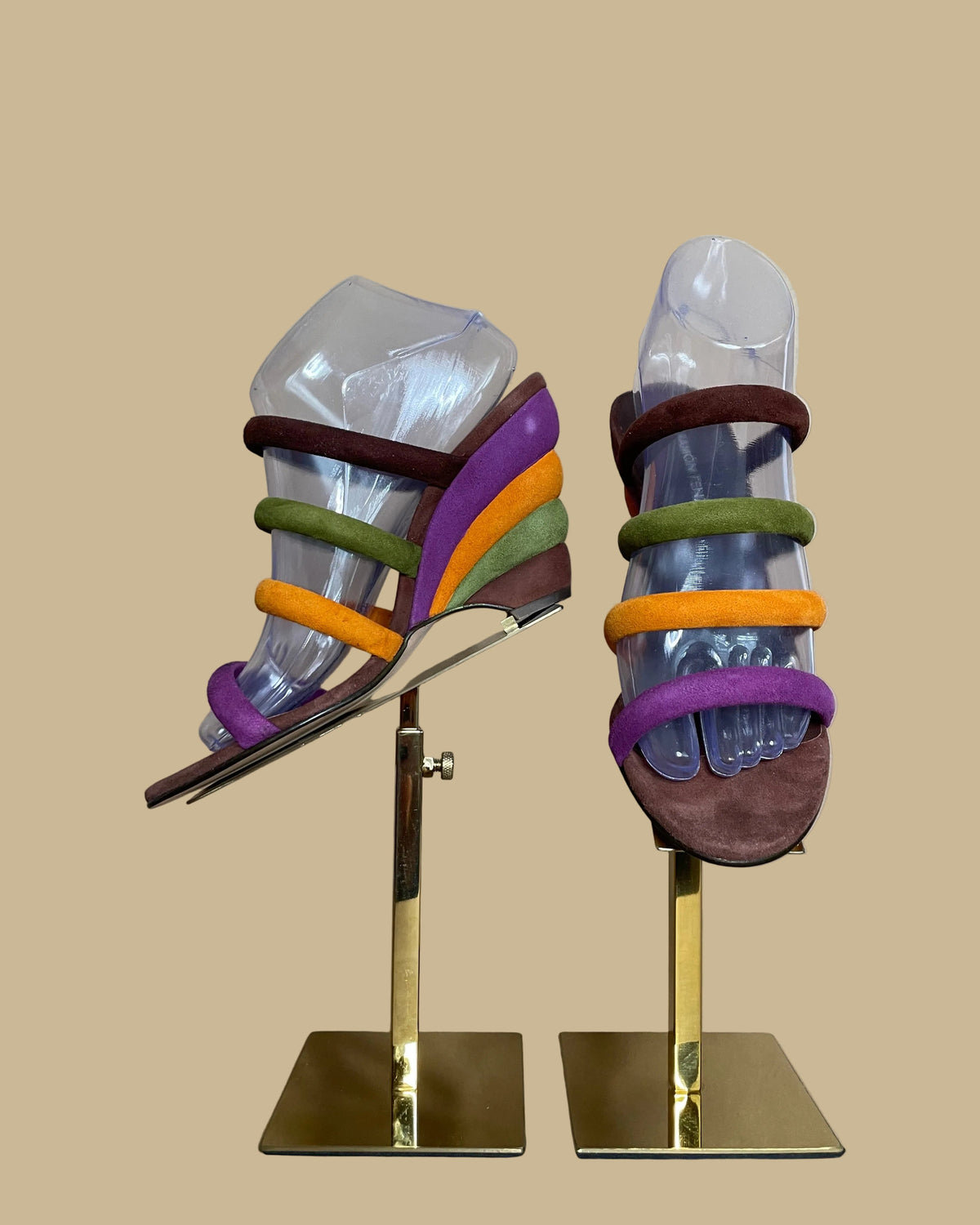 A pair of multi-strap suede wedge sandals in brown, olive, mustard, and purple, shown on clear display forms against a neutral beige background