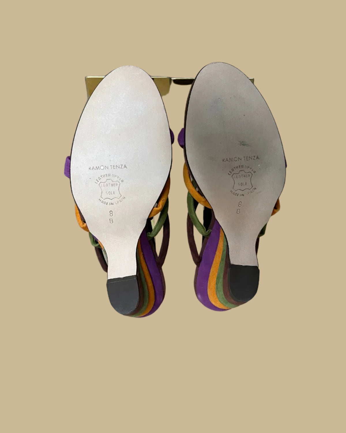 A pair of multi-strap suede wedge sandals in brown, olive, mustard, and purple, shown on clear display forms against a neutral beige background