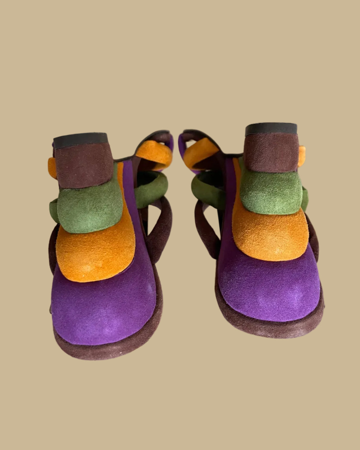 A pair of multi-strap suede wedge sandals in brown, olive, mustard, and purple, shown on clear display forms against a neutral beige background