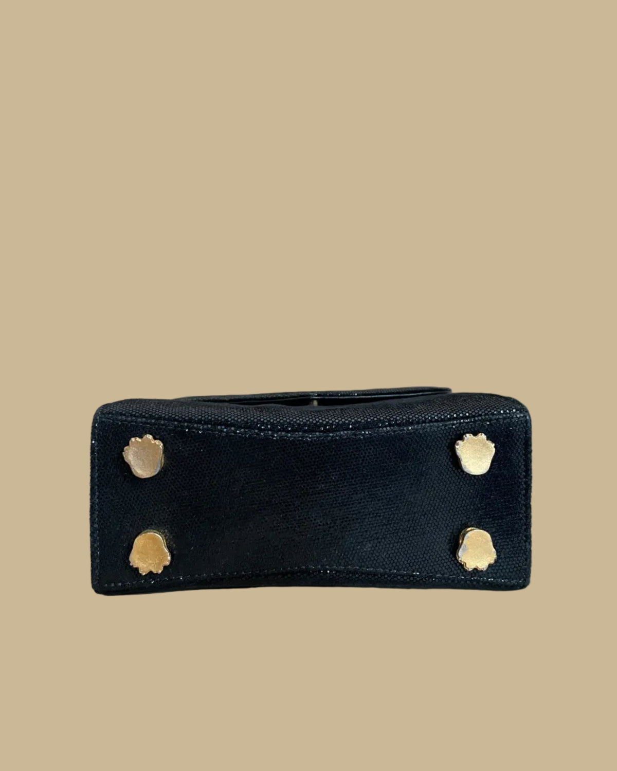 A small black textured handbag with a gold-tone chain strap and decorative gold-tone hardware, photographed against a neutral beige backdrop.