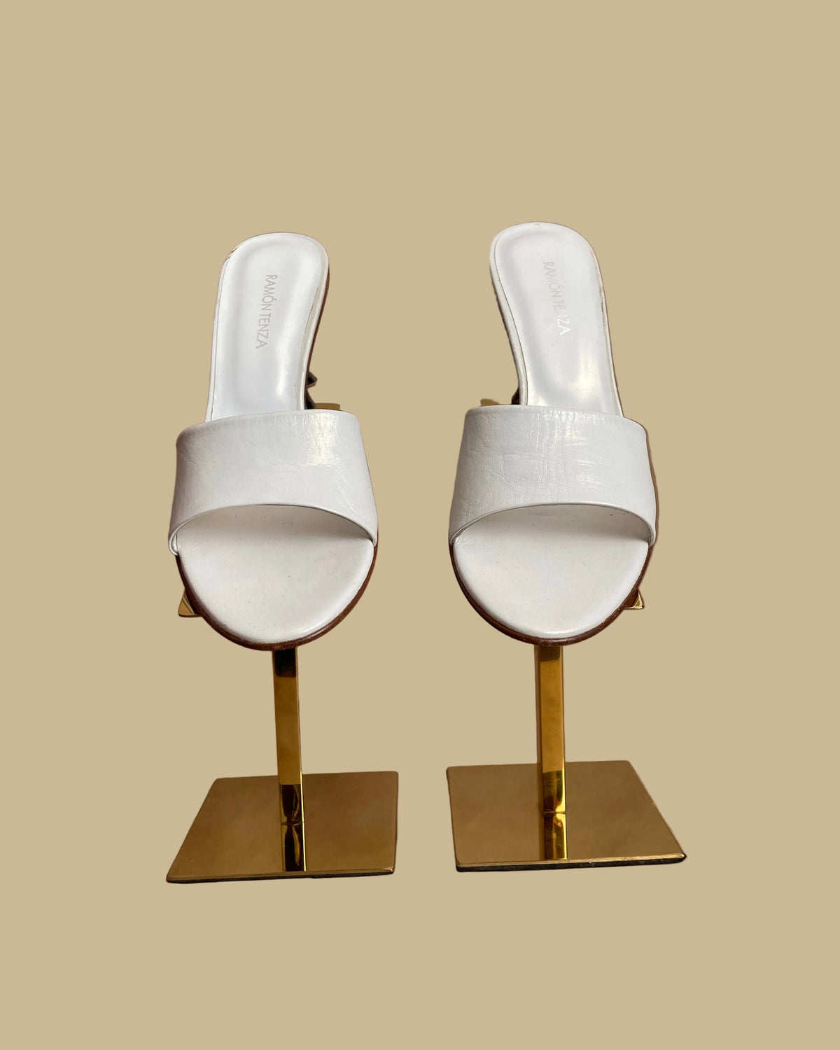 A pair of white leather Ramón Tenza slide sandals with a sculptural gunmetal elephant-shaped heel, displayed on gold stands against a kraft background