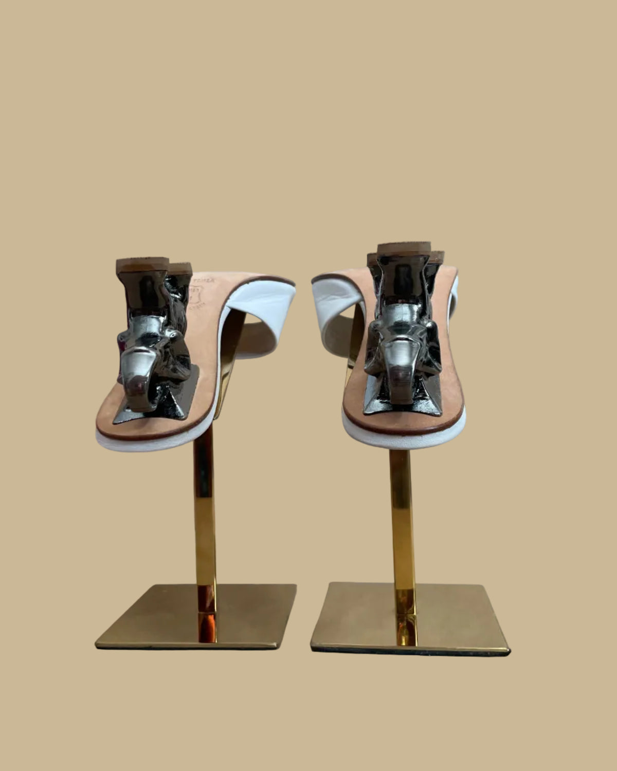 A pair of white leather Ramón Tenza slide sandals with a sculptural gunmetal elephant-shaped heel, displayed on gold stands against a kraft background