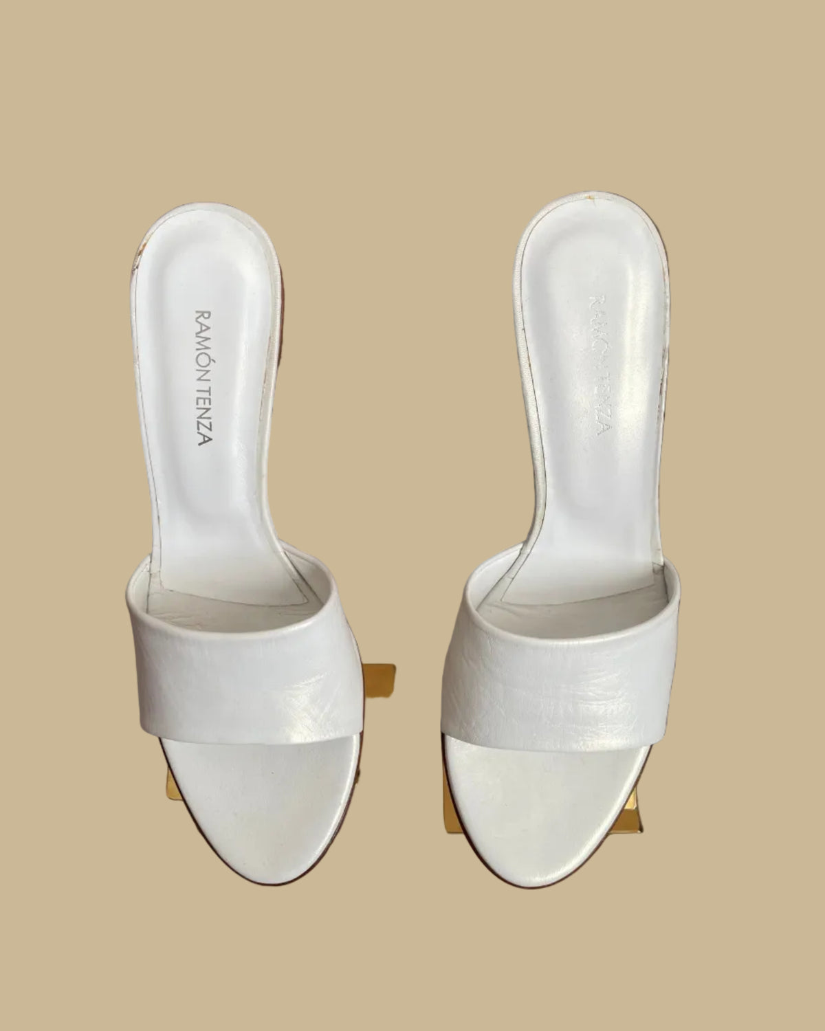 A pair of white leather Ramón Tenza slide sandals with a sculptural gunmetal elephant-shaped heel, displayed on gold stands against a kraft background