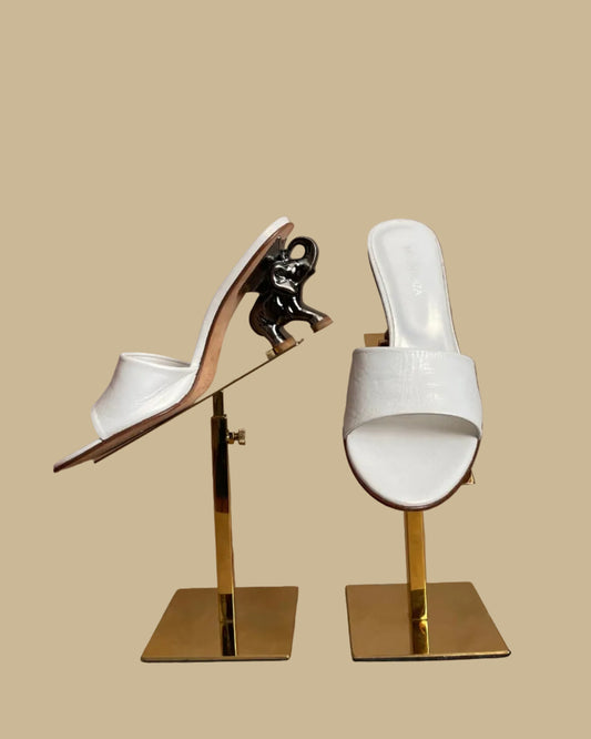 A pair of white leather Ramón Tenza slide sandals with a sculptural gunmetal elephant-shaped heel, displayed on gold stands against a kraft background