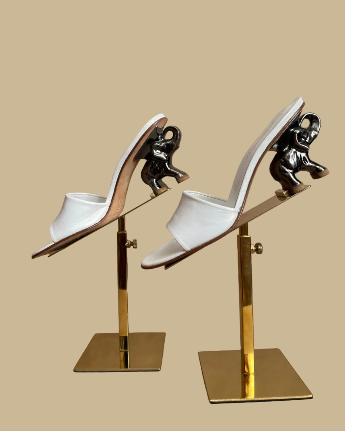 A pair of white leather Ramón Tenza slide sandals with a sculptural gunmetal elephant-shaped heel, displayed on gold stands against a kraft background