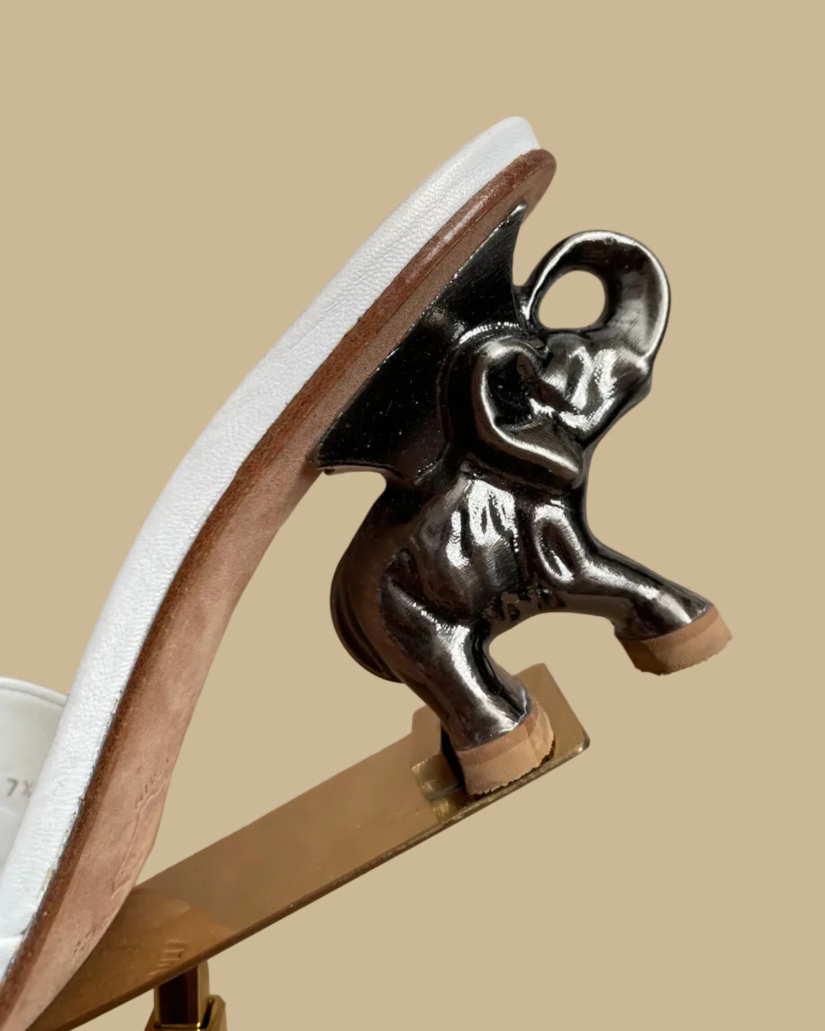 A pair of white leather Ramón Tenza slide sandals with a sculptural gunmetal elephant-shaped heel, displayed on gold stands against a kraft background