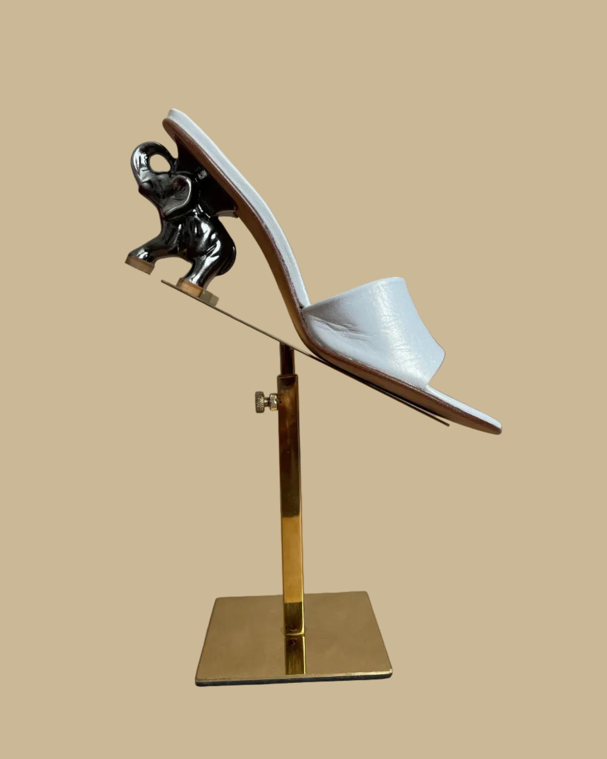 A pair of white leather Ramón Tenza slide sandals with a sculptural gunmetal elephant-shaped heel, displayed on gold stands against a kraft background
