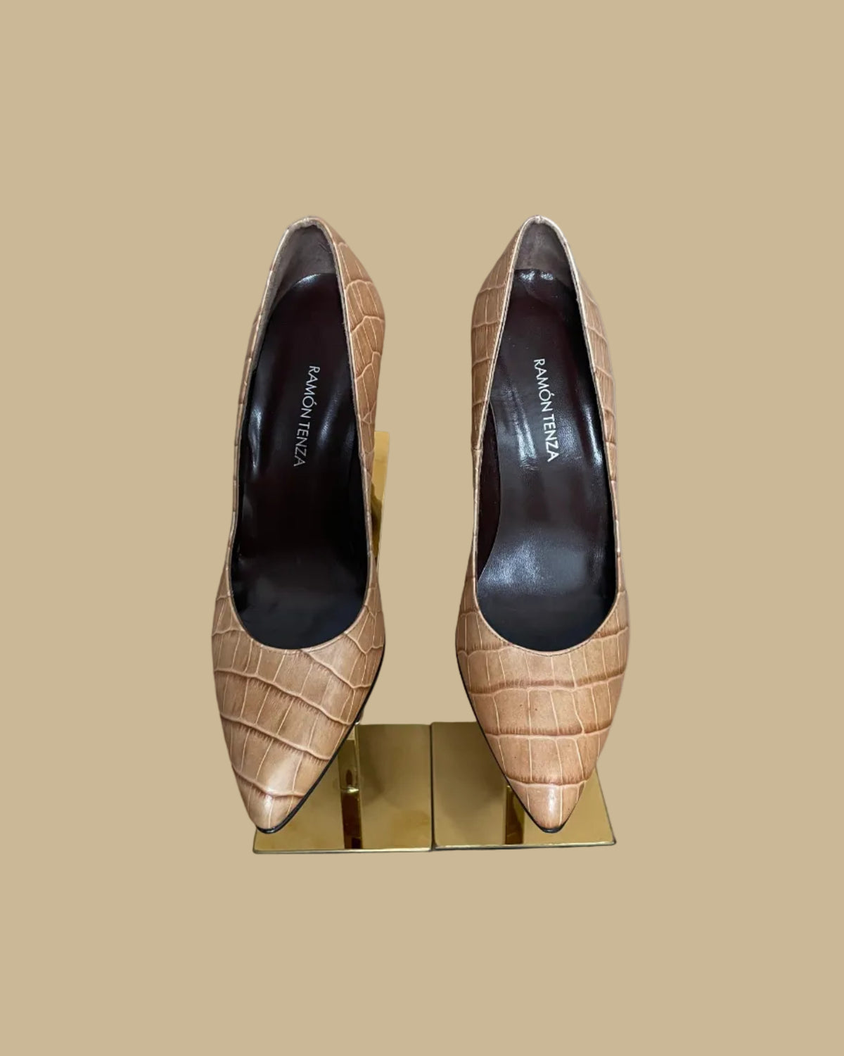 A pair of pointed-toe high heels in a tan crocodile-embossed leather, displayed on a gold-colored stand against a neutral beige background. The brand name Ramon Tenza is visible on the black insoles.