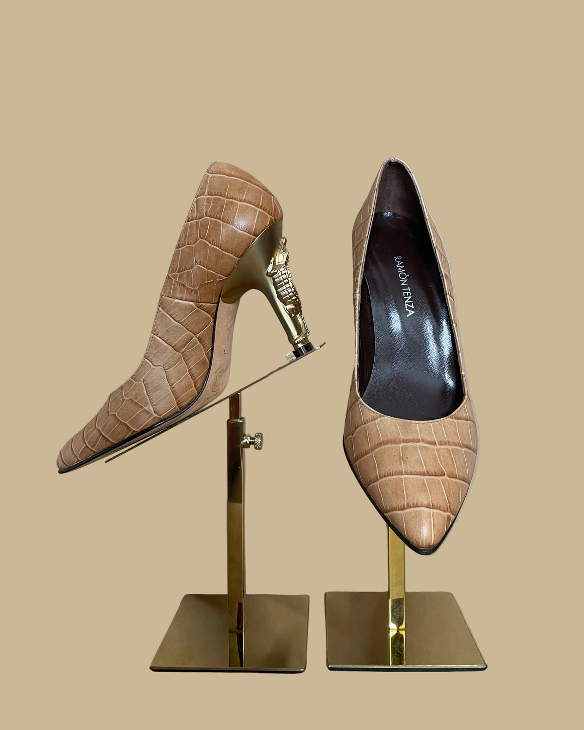 A pair of pointed-toe high heels in a tan crocodile-embossed leather, displayed on a gold-colored stand against a neutral beige background. The brand name Ramon Tenza is visible on the black insoles.