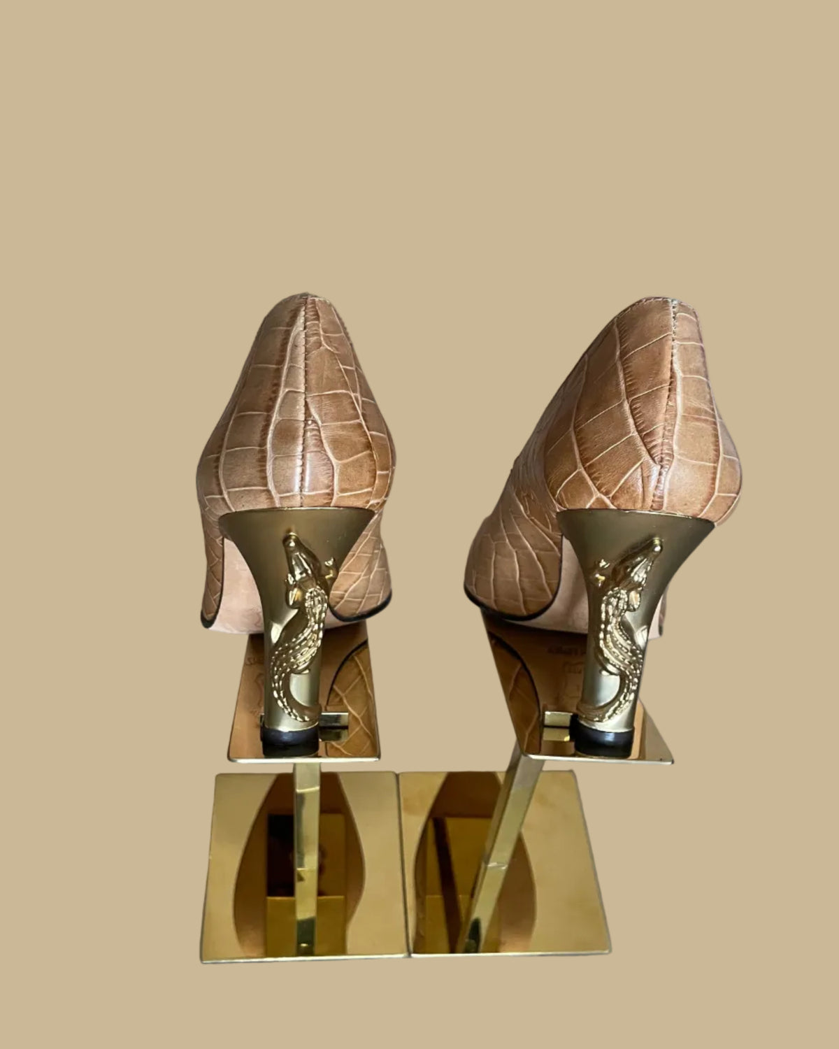 A pair of pointed-toe high heels in a tan crocodile-embossed leather, displayed on a gold-colored stand against a neutral beige background. The brand name Ramon Tenza is visible on the black insoles.