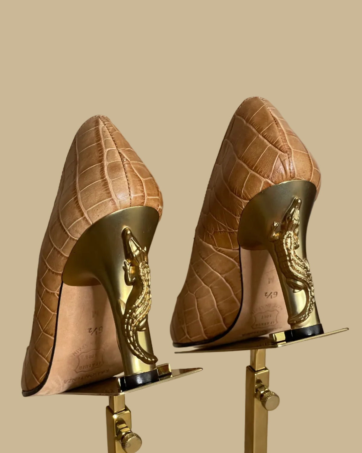 A pair of pointed-toe high heels in a tan crocodile-embossed leather, displayed on a gold-colored stand against a neutral beige background. The brand name Ramon Tenza is visible on the black insoles.