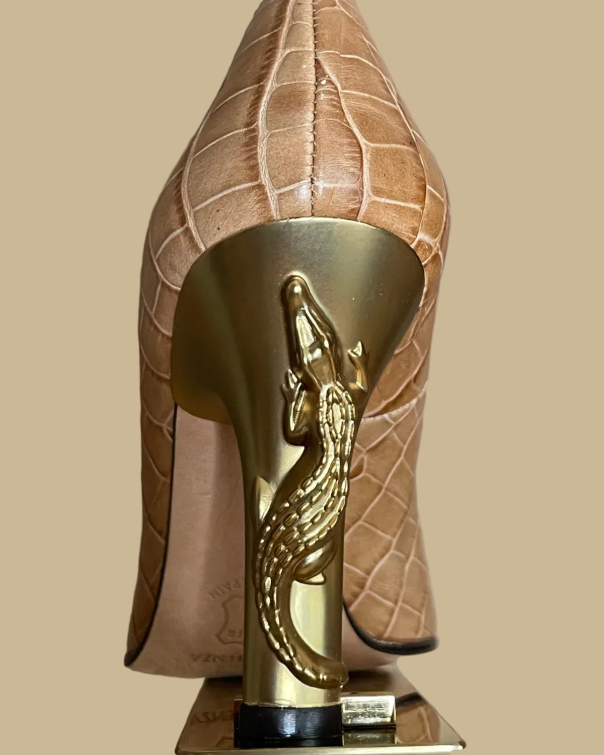 A close-up of a high-heeled shoe shown from the back, featuring a tan crocodile-embossed leather upper and a prominent gold heel with a sculpted alligator detail.